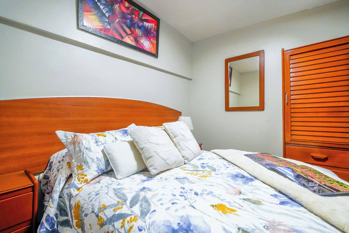 Quarto com cabeceira de madeira, roupa de cama floral, mesa de cabeceira, aparador, espelho e guarda-roupa.
