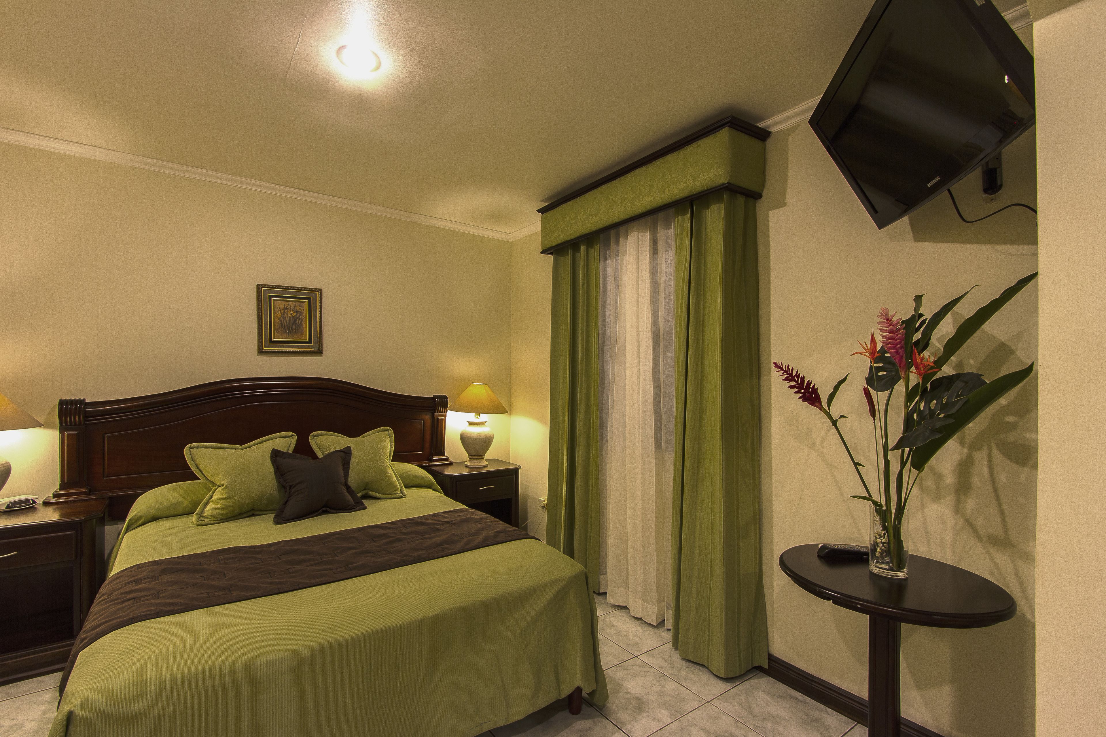 A hotel room with a neatly made bed, two bedside lamps, a small table with a vase of flowers, and a television mounted on the wall.