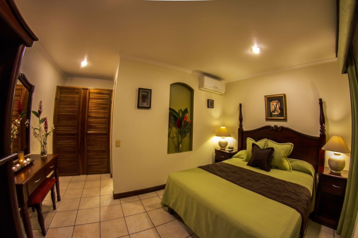 Habitación de hotel con cama, lámparas de noche, mesita de noche, escritorio con lámpara, espejo y puerta.