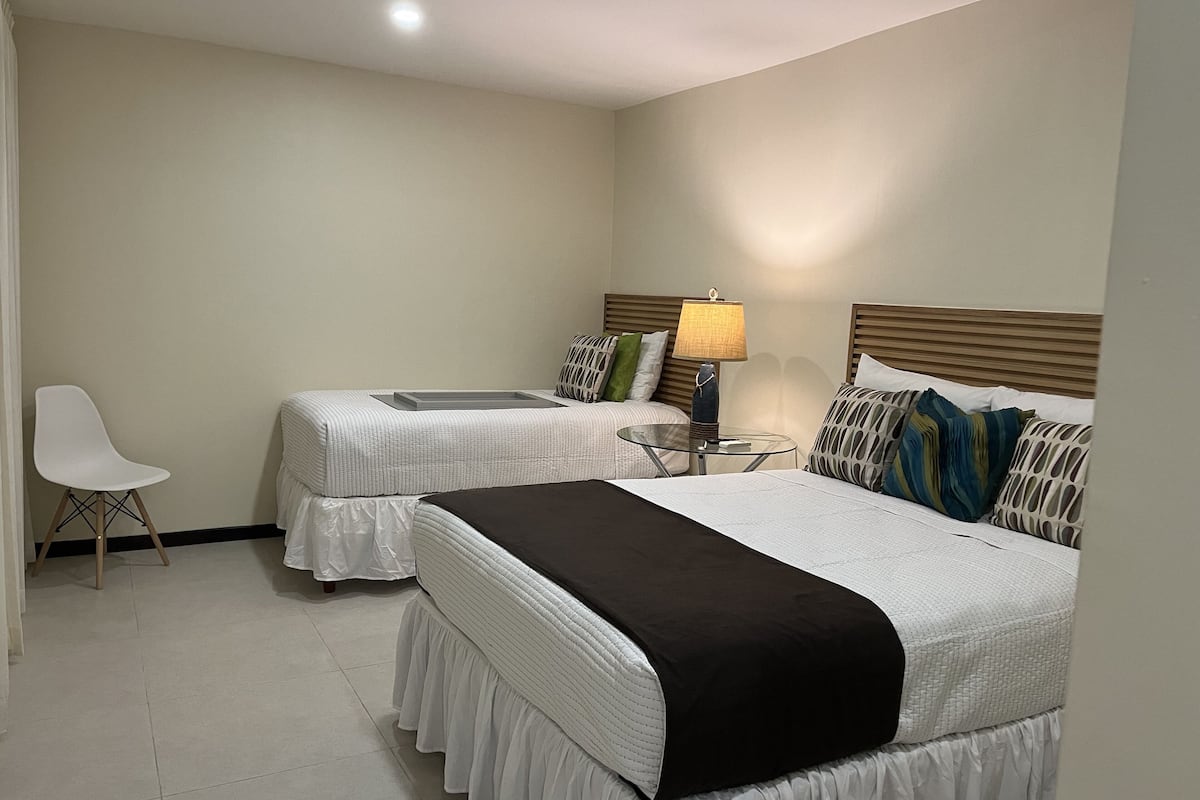 A hotel room with two beds, a chair, a bedside table with a lamp, and a small table between the beds.