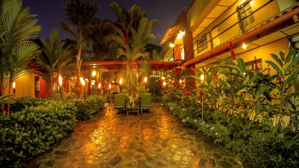A courtyard with stone flooring, palm trees, and a building with a balcony. - Hotel Boutique Iguana Verde (Orotina)