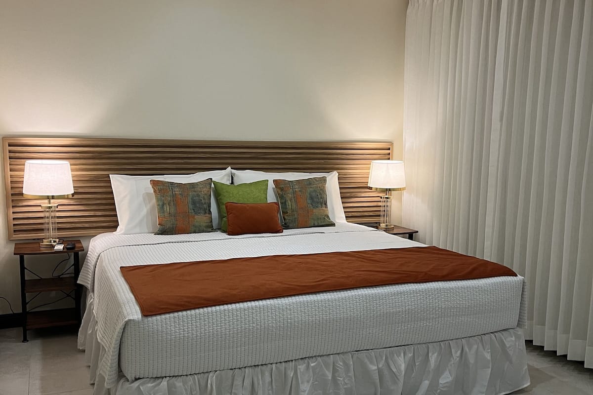 A hotel room with a large bed, two bedside tables with lamps, a wooden headboard, and a view of a curtain.