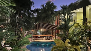 A pool area with a pergola, surrounded by tropical plants and a building with yellow walls. - Hotel Boutique Iguana Verde (Orotina)