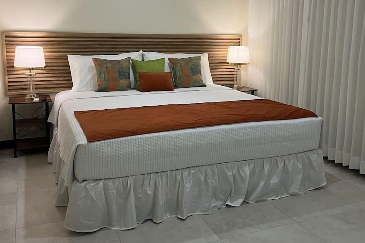 Una habitación de hotel con una cama grande, dos lámparas de noche, una mesita de noche y un ventanal con cortinas.