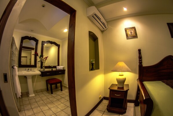 A bathroom with a sink, mirror, vanity, and a bedside table with a lamp.