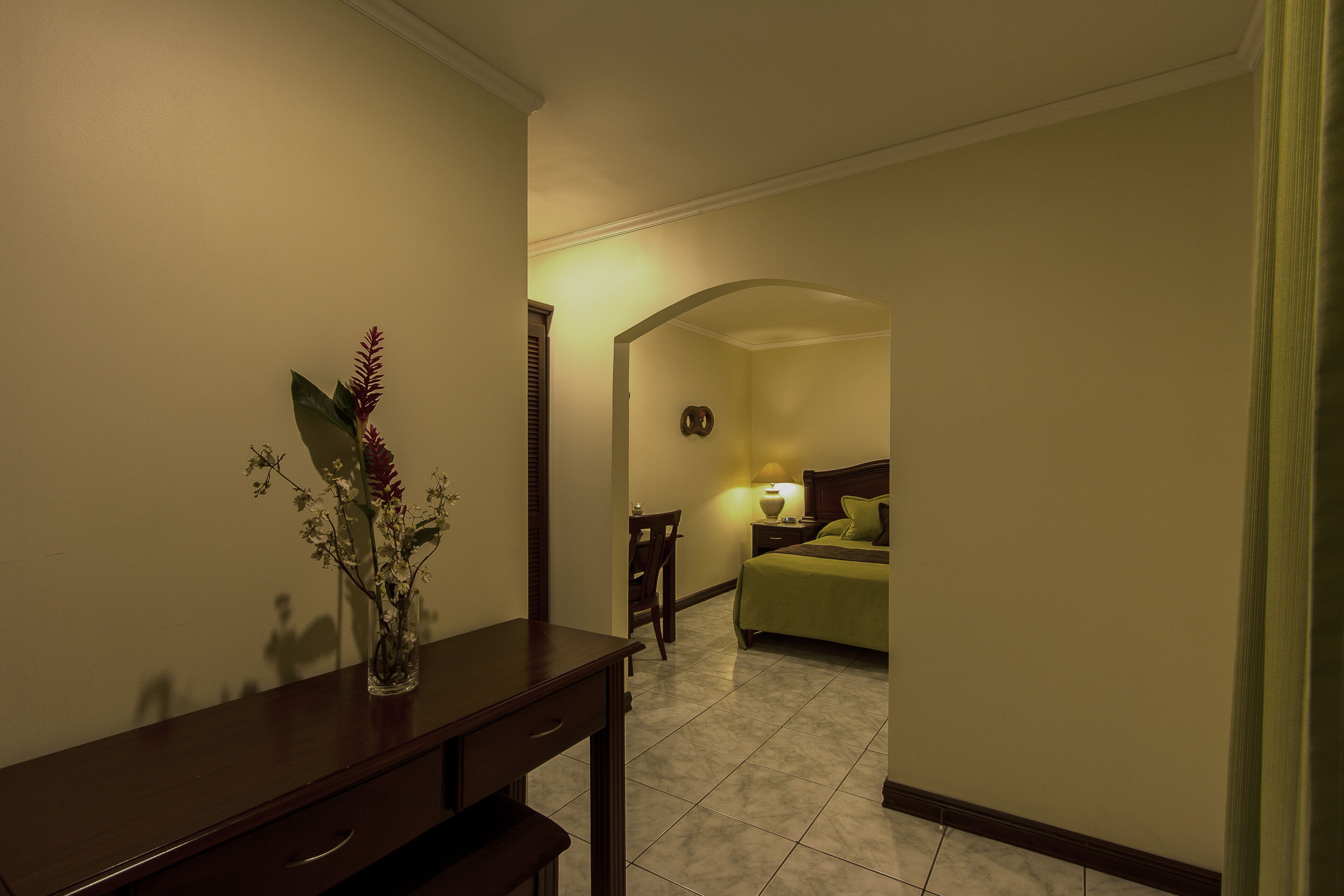 A hotel room with a bed, bedside table, lamp, and a desk with a vase of flowers.