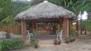 A thatched-roof gazebo with brick pillars, surrounded by potted plants and white plastic chairs. - Pousada do Riacho (Barreirinhas)