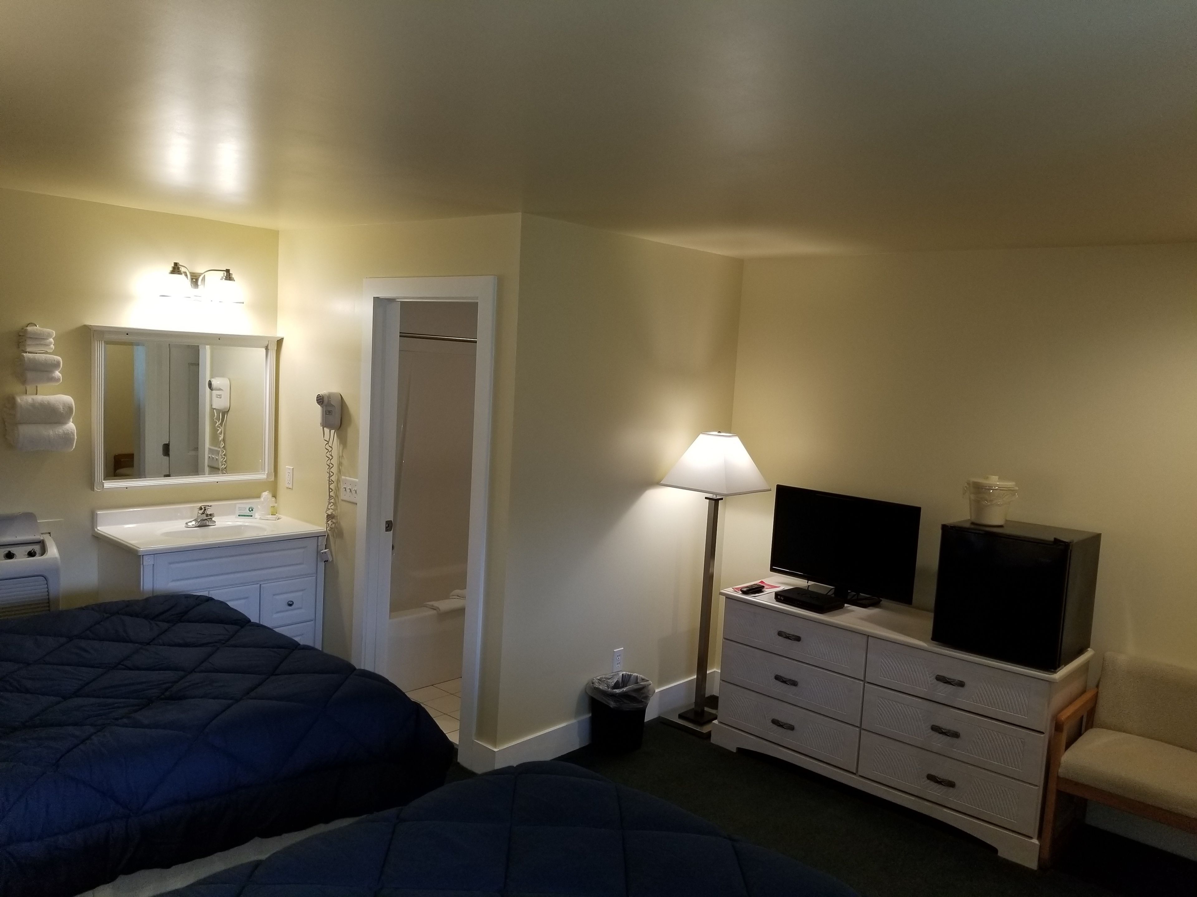 Quarto de hotel com duas camas, banheiro com espelho, pia, televisão e luminária.