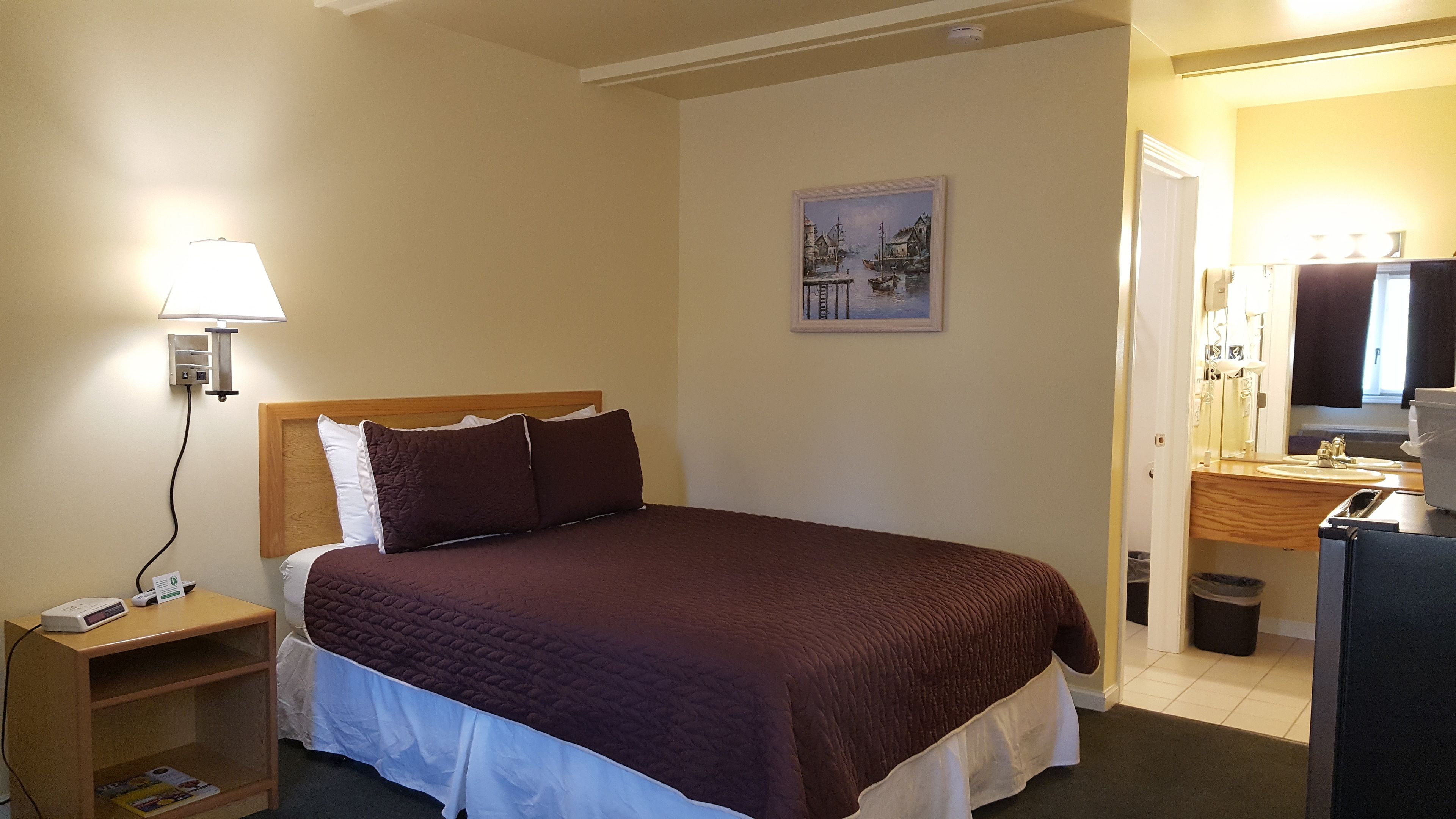 Een hotelkamer met een bed, nachtkastje, lamp, schilderij en een zichtbare badkamer door een open deur.