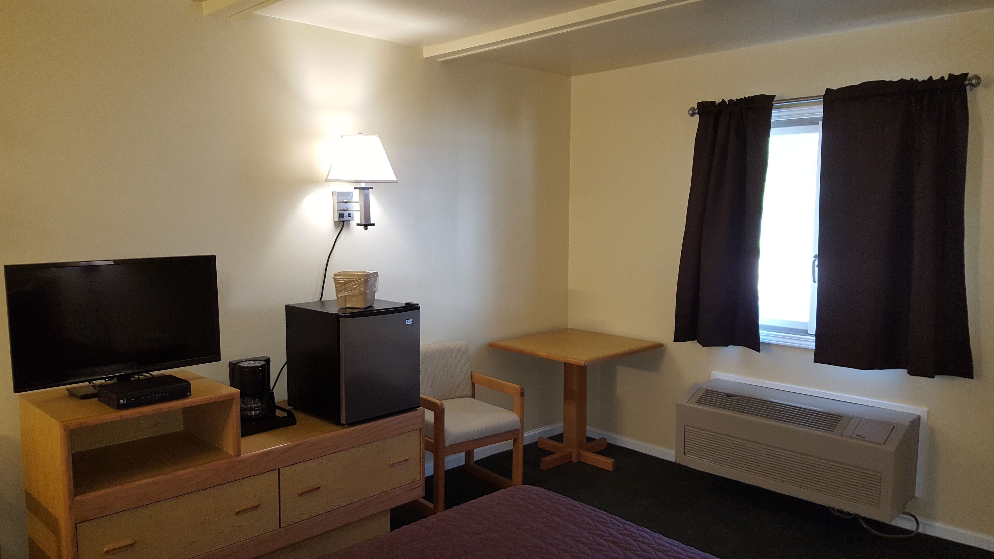 A hotel room with a TV, a desk, a chair, a bed, a heater, and a window with curtains.