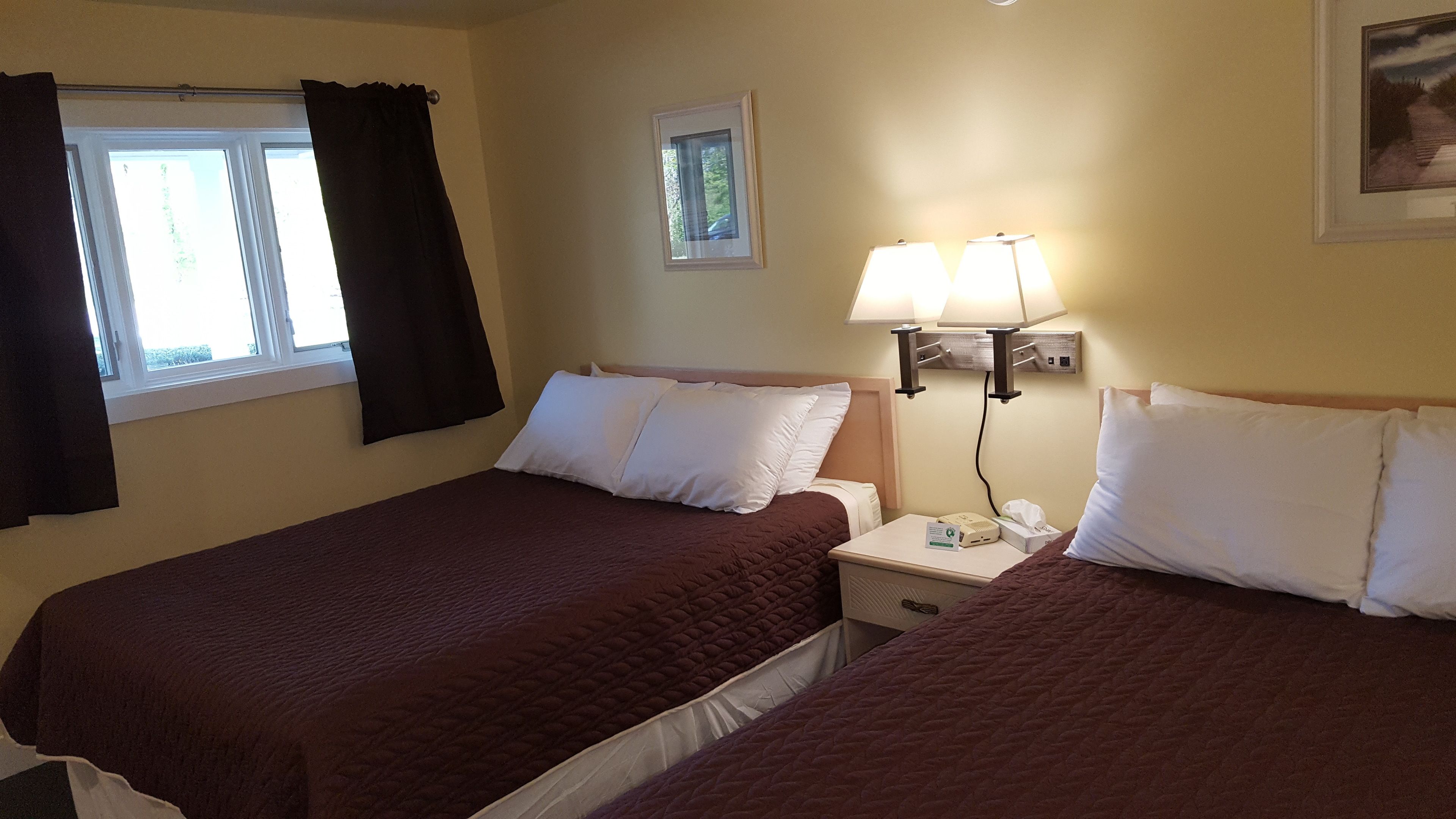 A hotel room with two beds, a window with curtains, a bedside lamp, and a small picture on the wall.