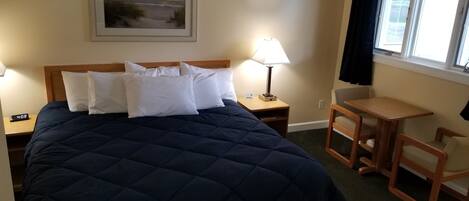 Une chambre d’hôtel avec un grand lit, une table de chevet avec une lampe, une petite table en bois et une chaise. Il y a un tableau au mur et une fenêtre avec des rideaux.
