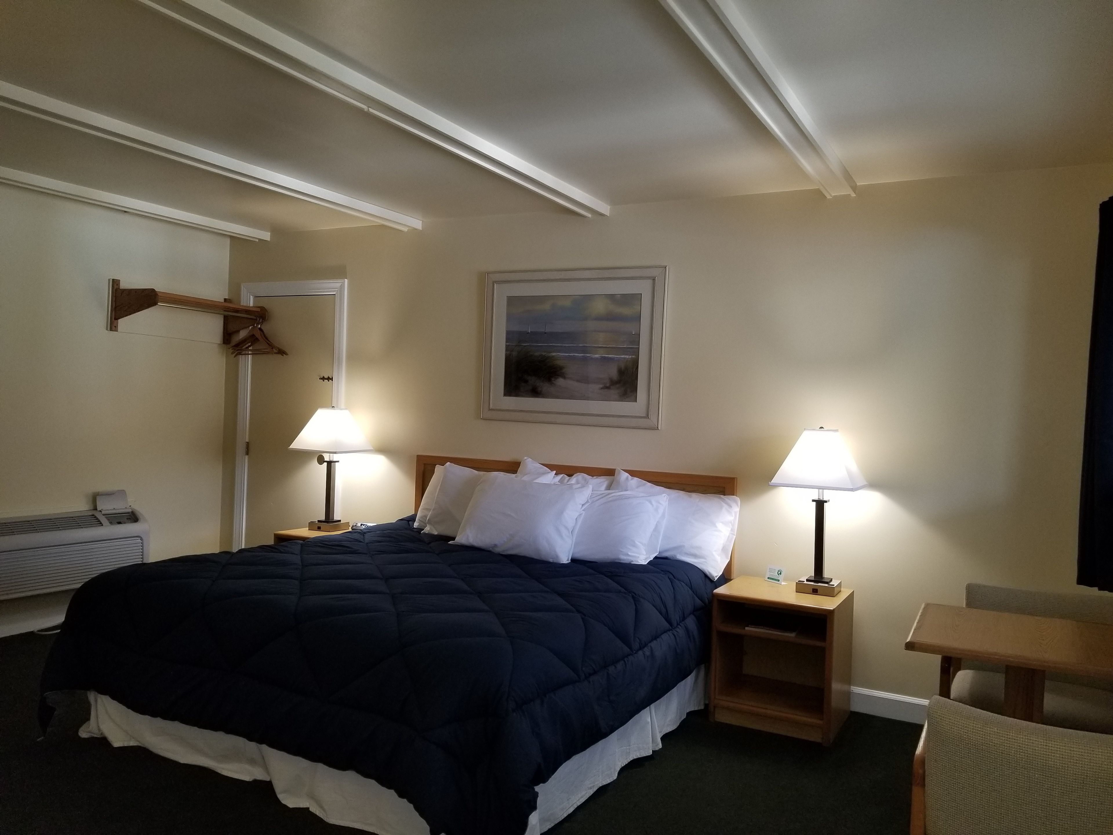 A hotel room with a bed, two bedside tables with lamps, a wooden chair, a radiator, and a framed picture on the wall.