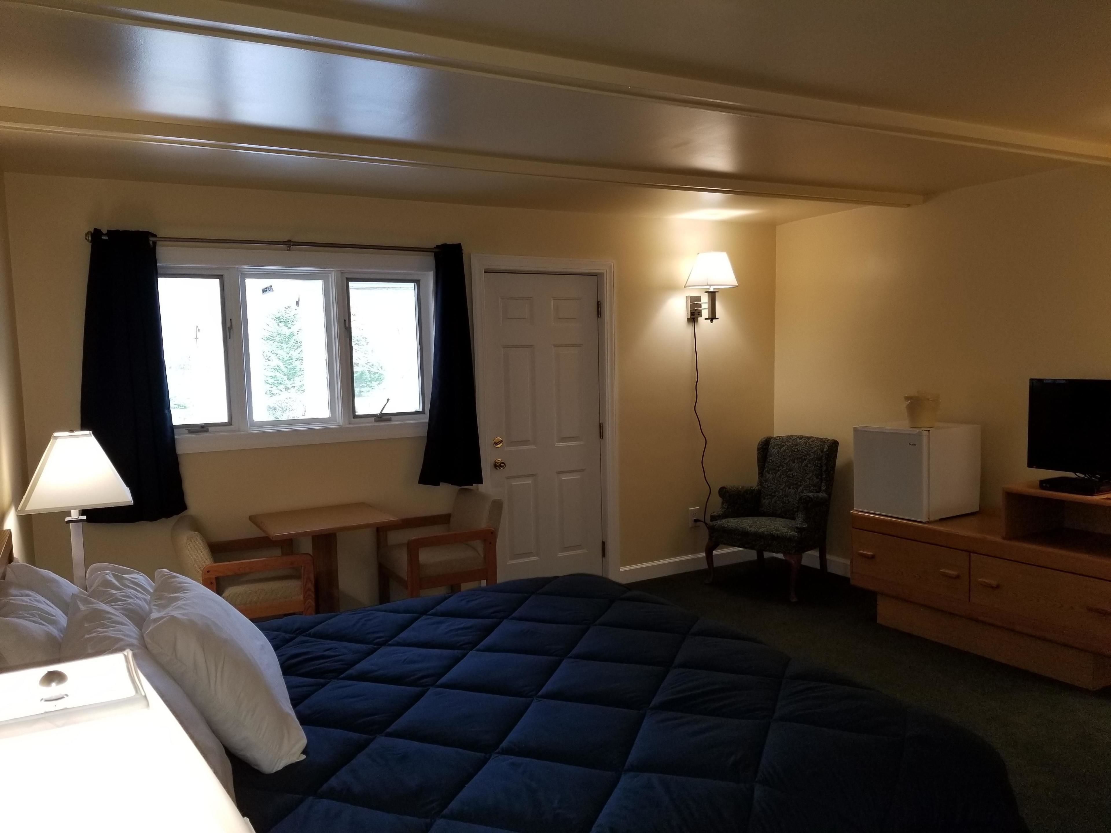 Quarto de hotel com cama, escrivaninha, cadeira, televisão e luminária.