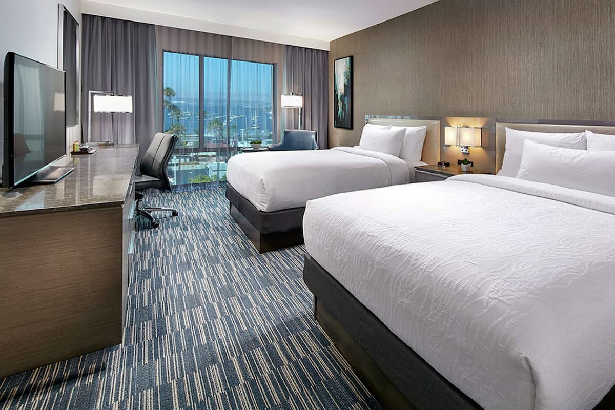 Habitación de hotel con dos camas, un escritorio con televisor, un separador de vidrio y vistas a la ciudad.