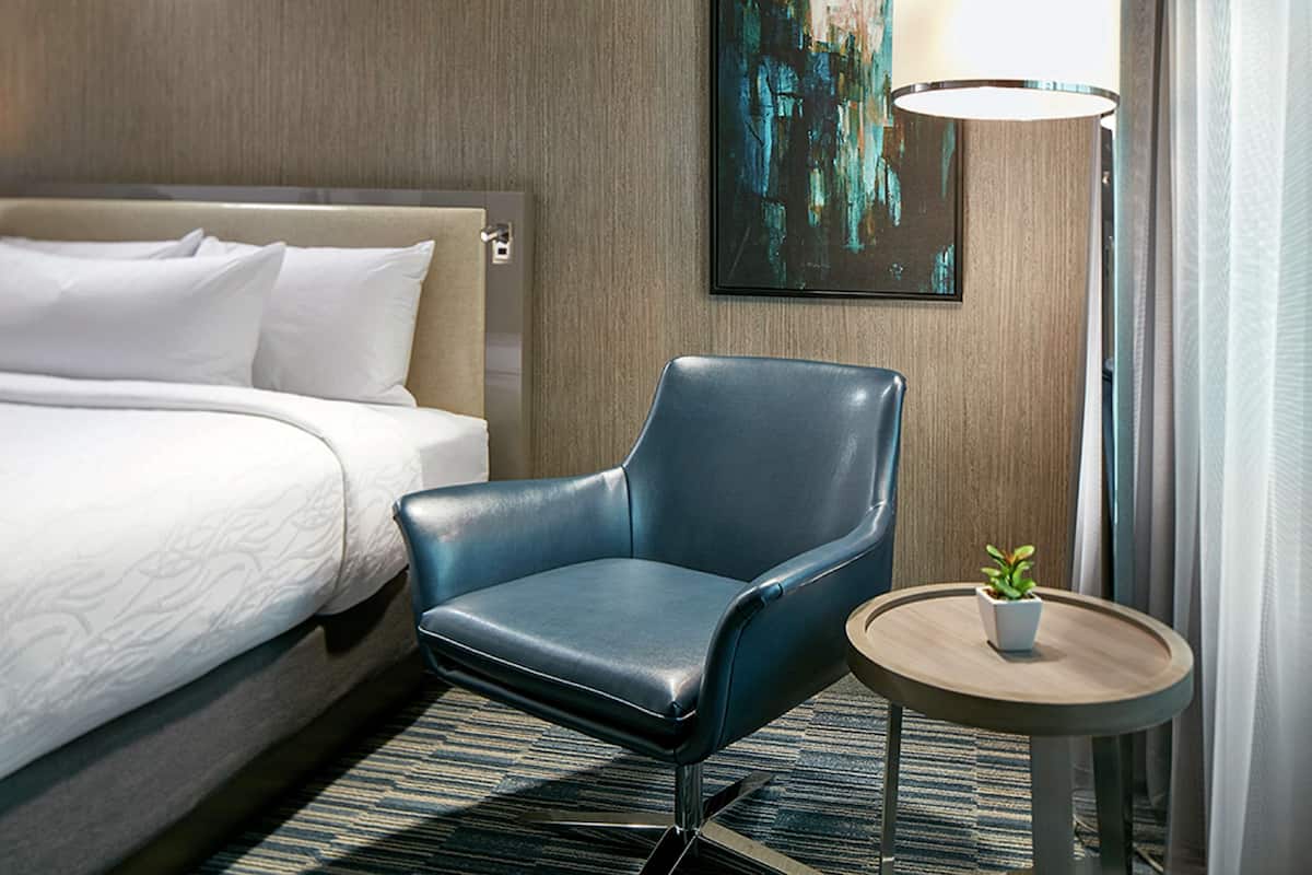 Una habitación de hotel con una cama, un sillón azul, una mesita redonda con una planta y un cuadro en la pared.