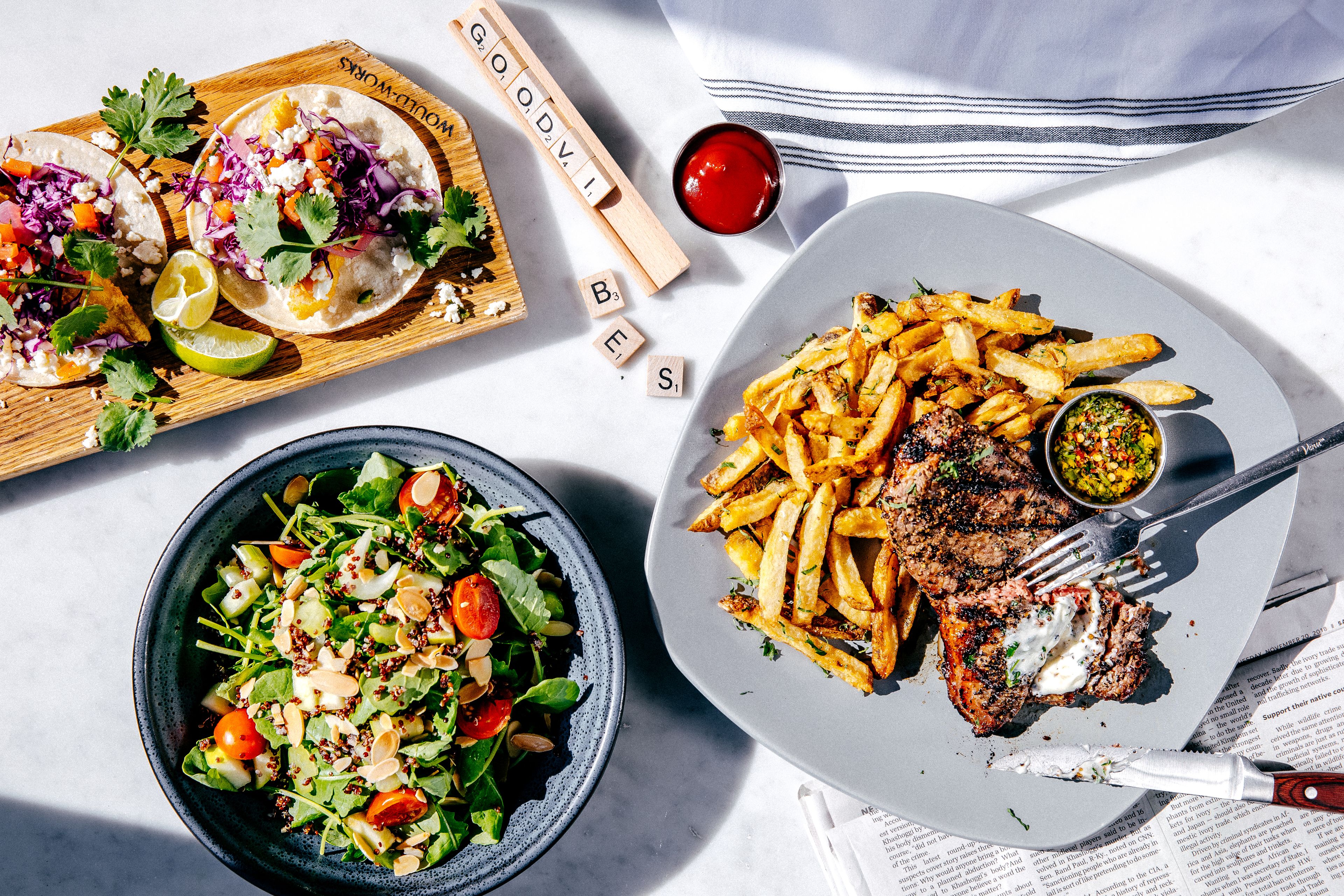 A table with a plate of steak and fries, a bowl of salad, and a wooden board with tacos.