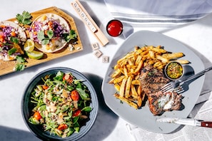 Una mesa con un plato de carne, papas fritas, un tazón de ensalada y una tabla de madera con tacos.