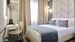 Quarto de hotel com cama, escrivaninha, cadeira, cabideiro e parede com padrão geométrico.