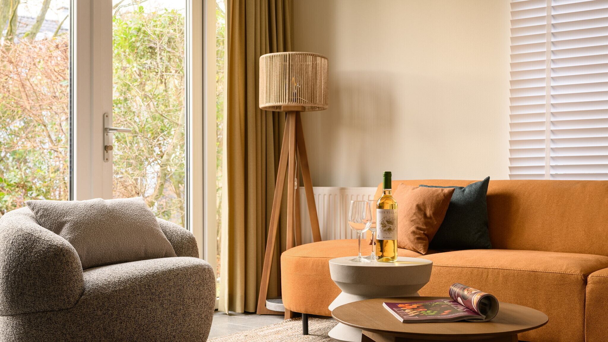 Een moderne woonkamer met een bank, fauteuil en een vloerlamp. Op een bijzettafeltje staan een fles wijn en een glas.