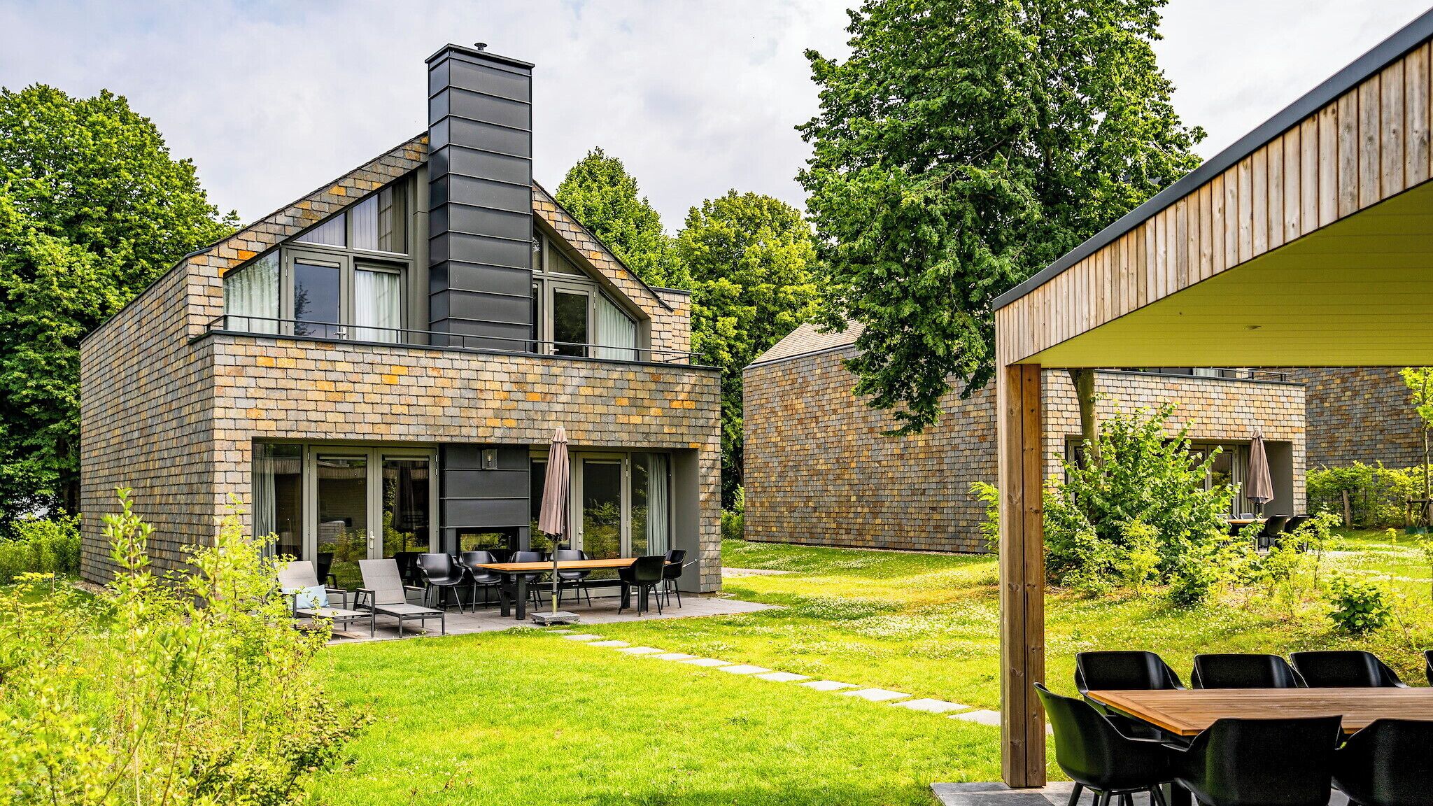 A modern house with a stone facade, a large chimney, and a covered outdoor seating area with a table and chairs.