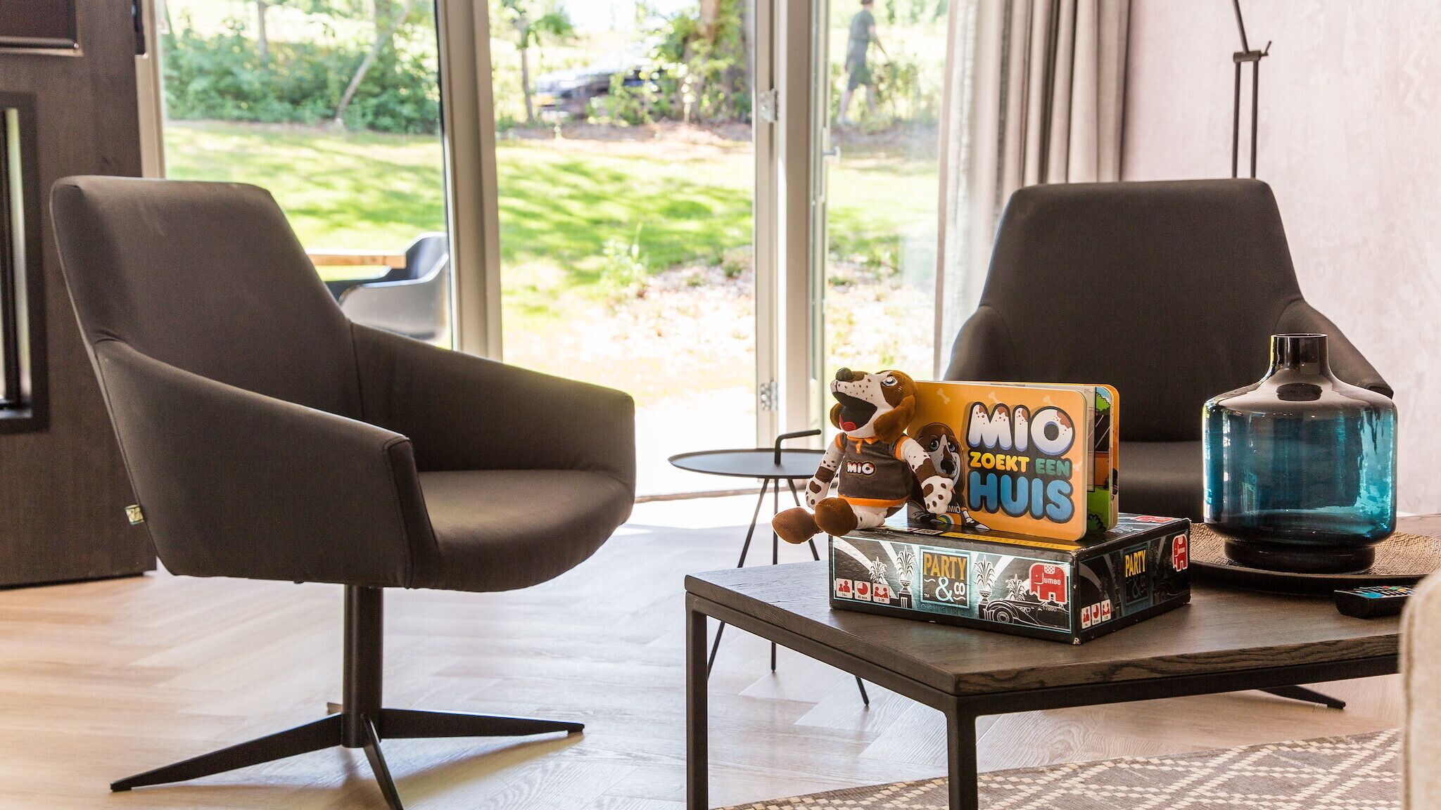 Ein modernes Wohnzimmer mit zwei dunkelgrauen Sesseln, einem Couchtisch und einem Gesellschaftsspiel mit dem Titel "MIO ZOEKT EEN HUIS".