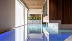 A modern indoor pool area with clear glass walls, a wooden ceiling, and a stone wall with plants. - Dormio Resort Maastricht (Maastricht)