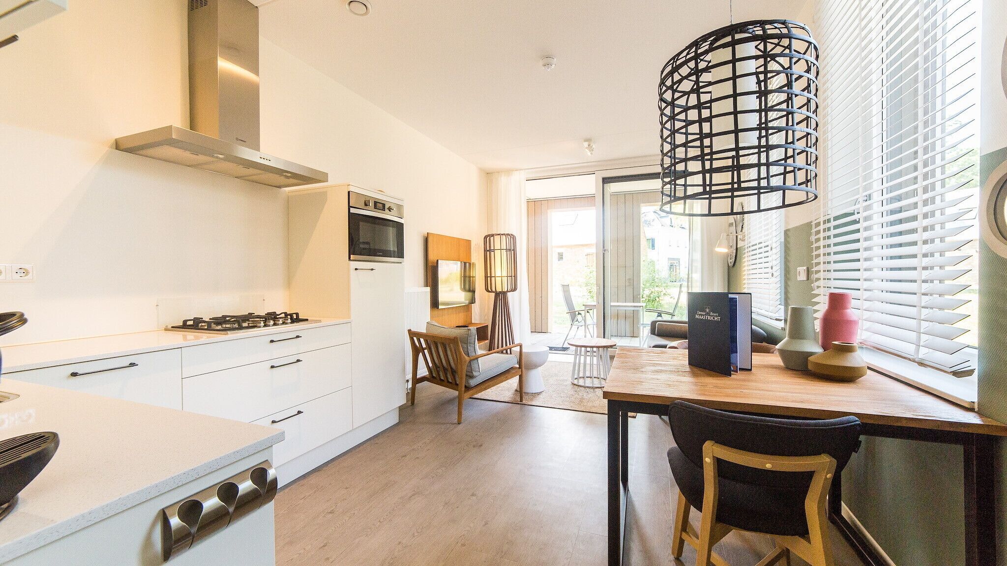 A modern kitchen with white cabinetry, a gas stove, and a built-in oven. There is a dining area with a wooden table and chairs, and a large window with blinds.