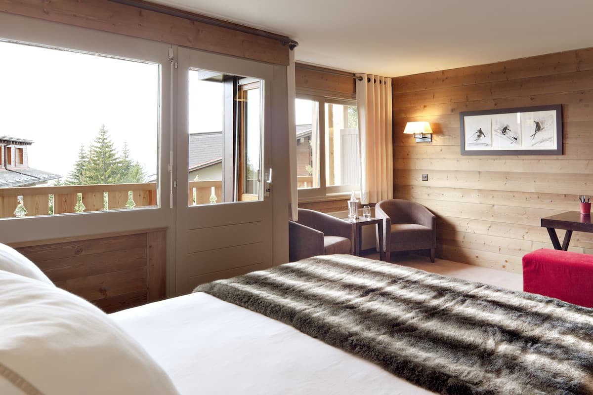 A hotel room with a bed, a desk with a chair, a small table, and a window with curtains.