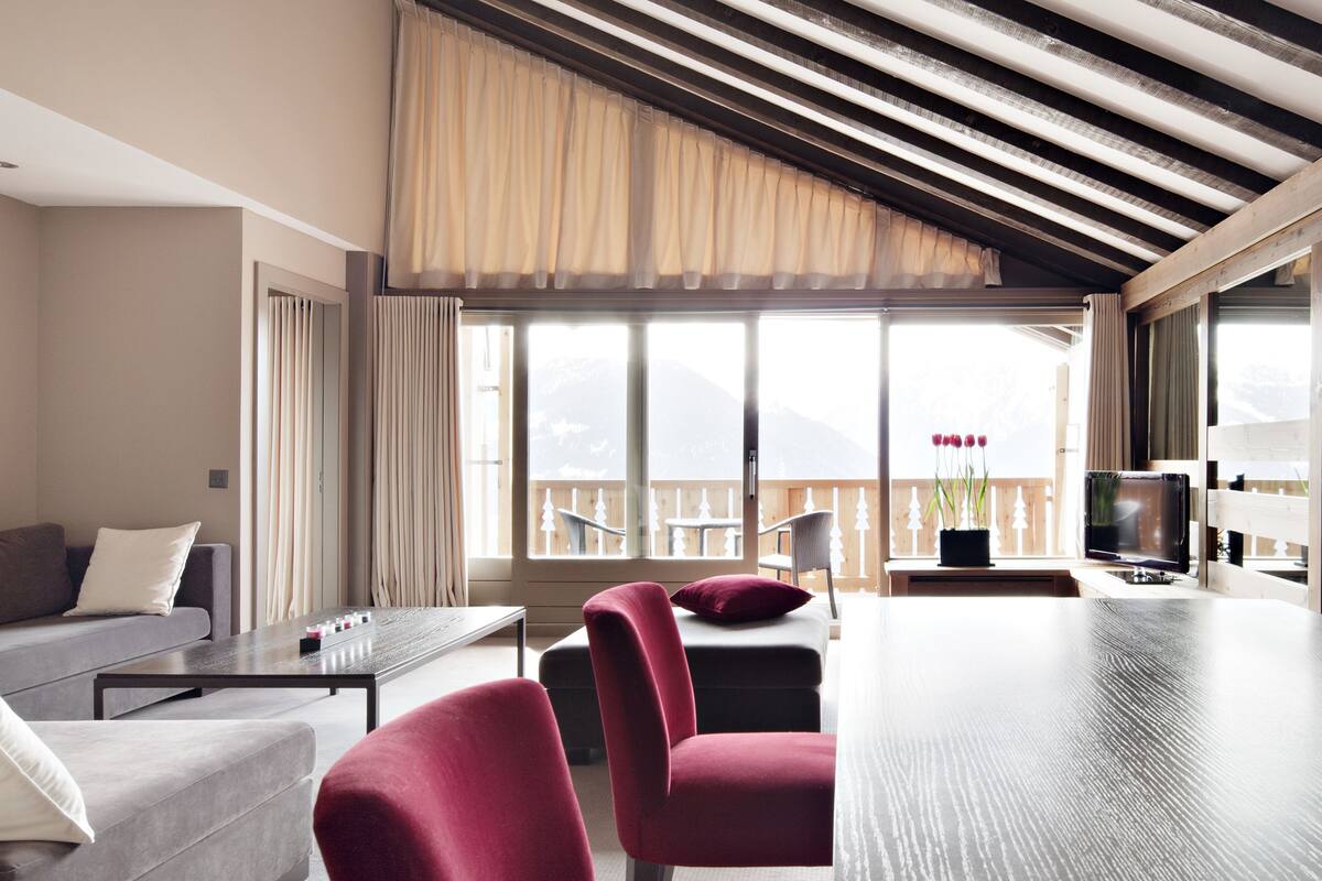 A modern living room with a wooden ceiling, large windows, a sofa, a dining table, and red chairs.