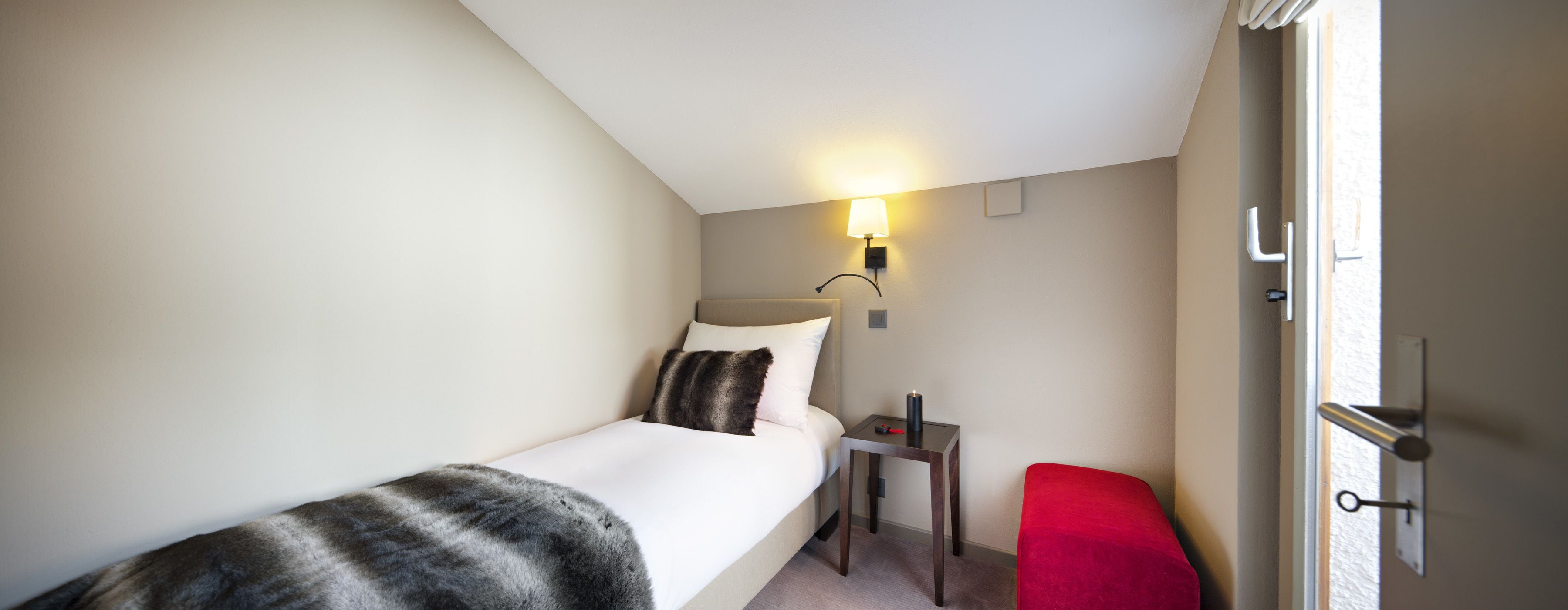 A small, neatly arranged bedroom with a single bed, a bedside table with a lamp, a red chair, and a door with a keyhole.