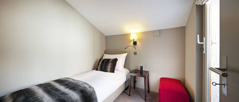 Un dormitorio pequeño y ordenado con una cama individual, una mesita de noche con una lámpara, una silla roja y una puerta con ojo de cerradura.
