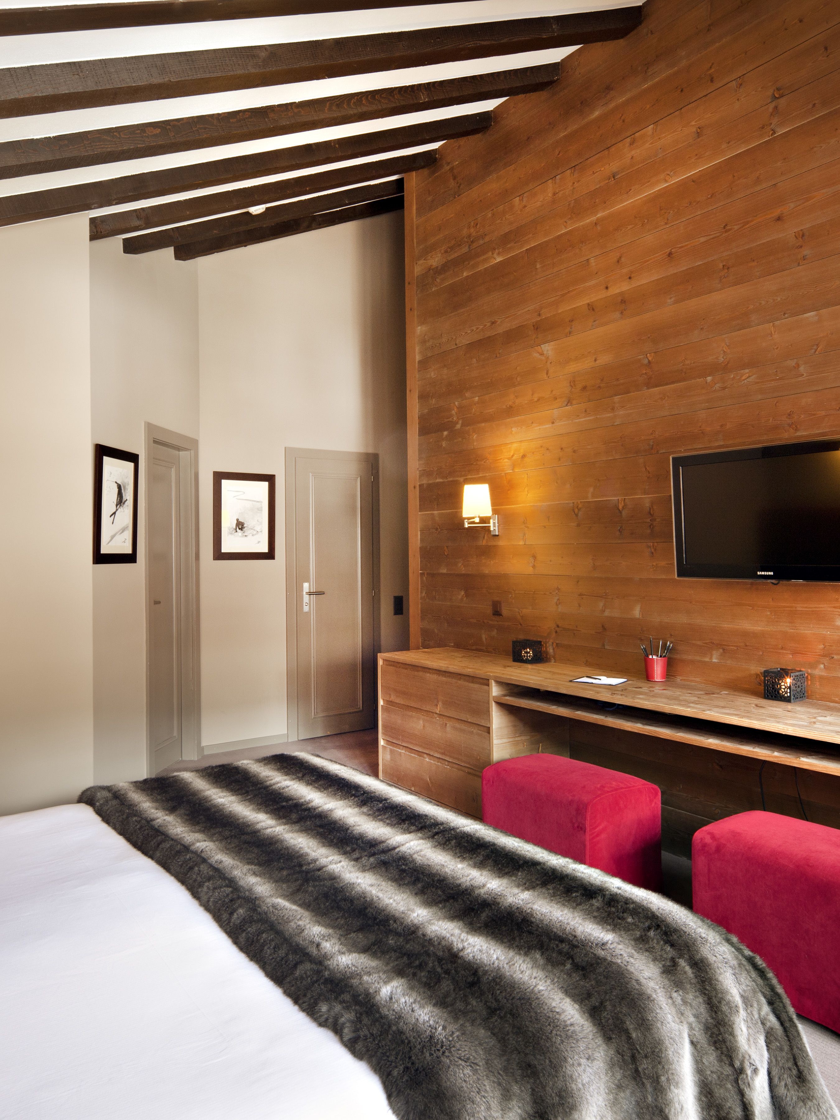 Una habitación de hotel moderna con techo de madera, un televisor de pantalla plana grande montado en la pared, un escritorio de madera con lámpara y una cama con manta a rayas.