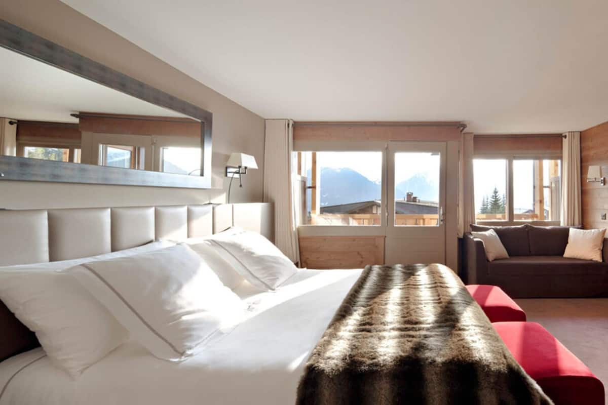 A hotel room with a bed, a red sofa, a wooden wall, and a window with a mountain view.