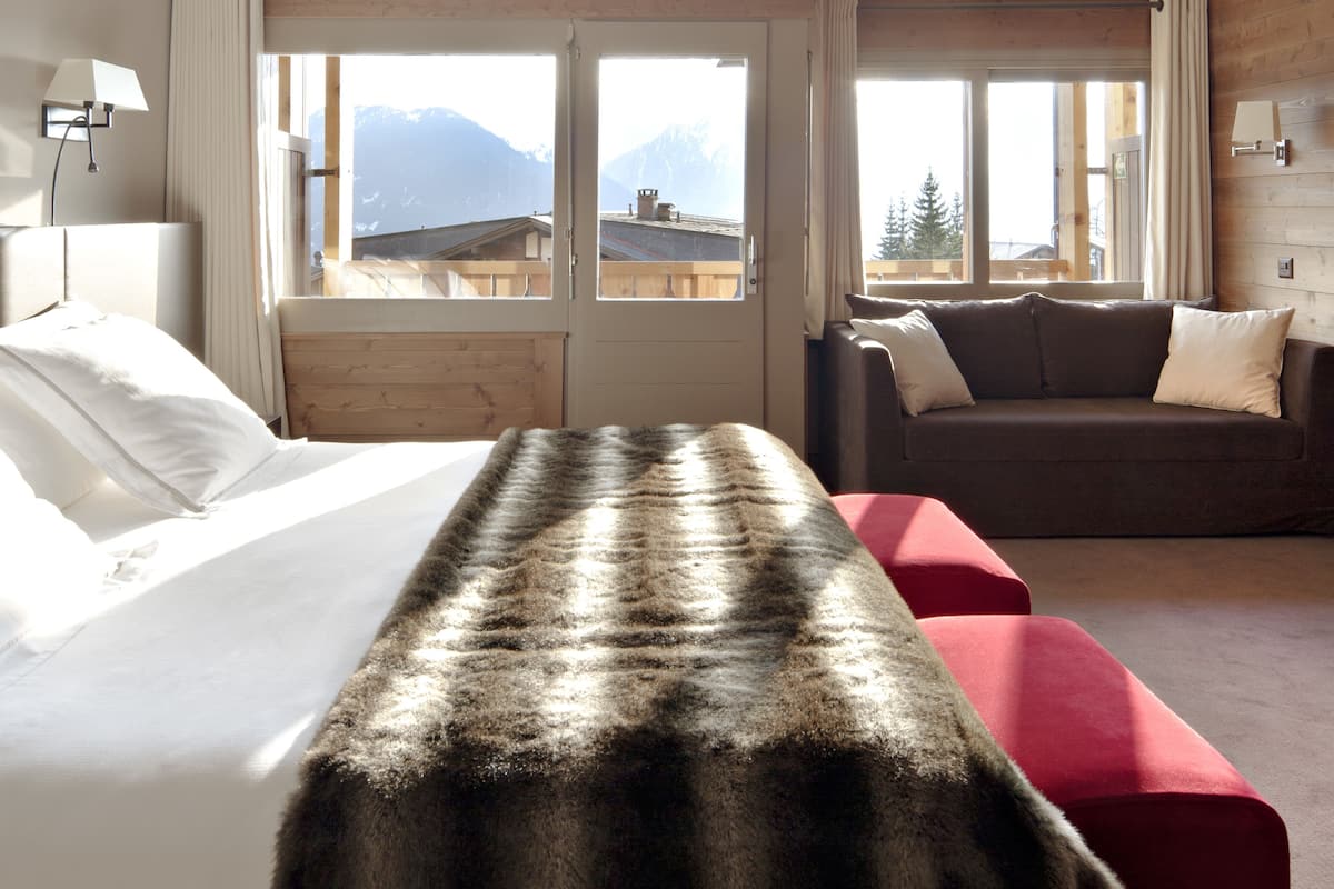 A hotel room with a bed, a sofa, and a red ottoman. There are windows with a mountain view.