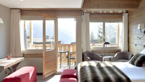 A room with a bed, a desk, and a chair, with a view of mountains through large windows.