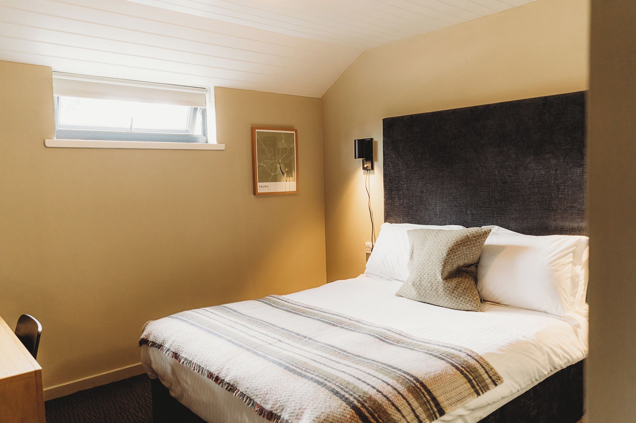 A bedroom with a bed, a bedside table, a window, and a wall-mounted light fixture.