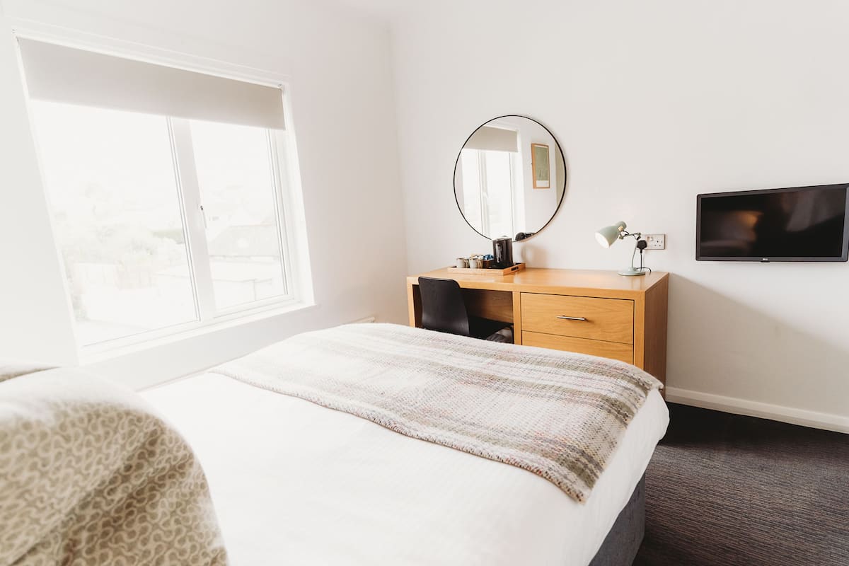 Ett hotellrum med en säng, ett skrivbord med en stol, en rund spegel, en platt-TV och ett fönster med persienner.