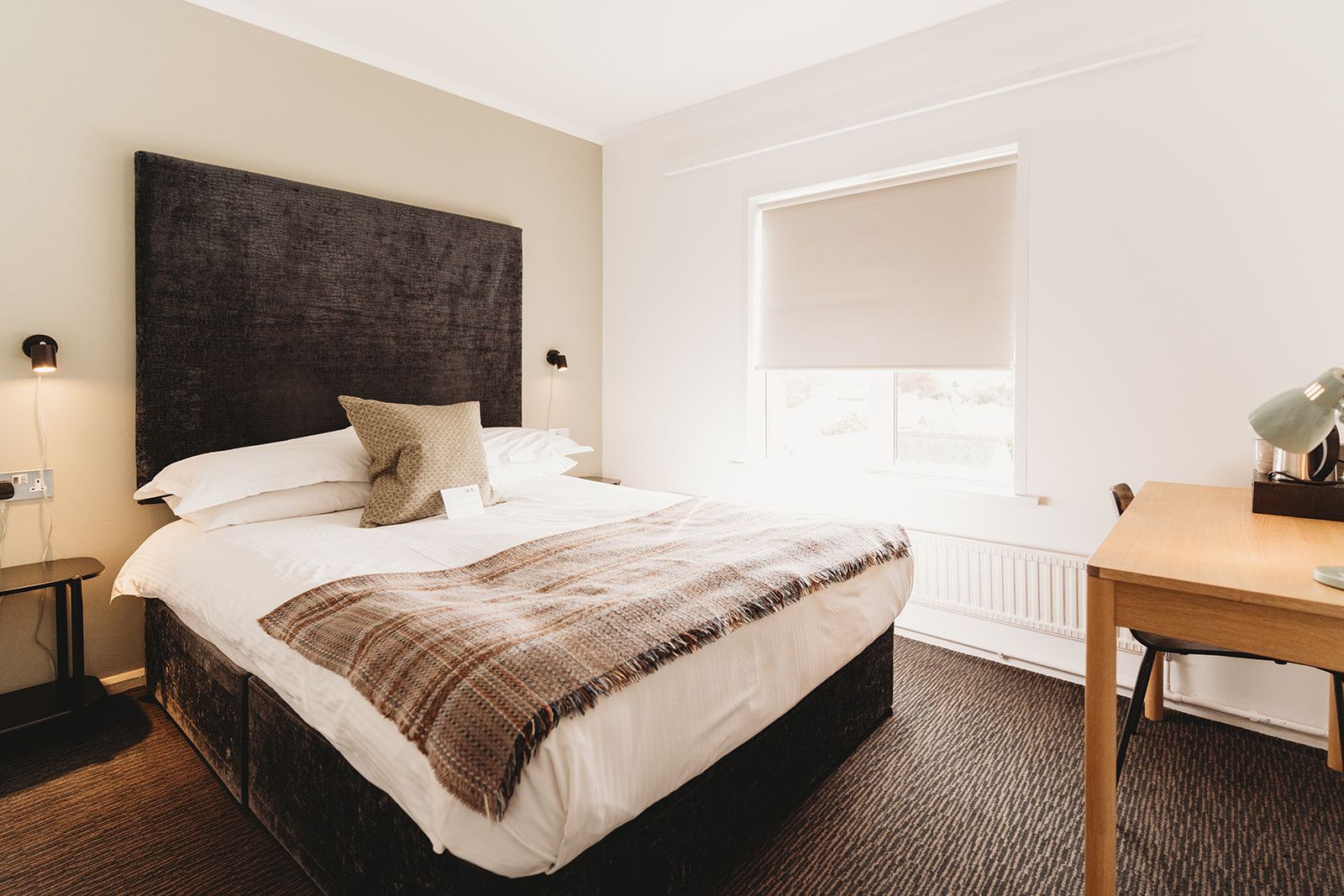 A hotel room with a large bed, bedside lamps, a window with blinds, and a small table with a lamp.