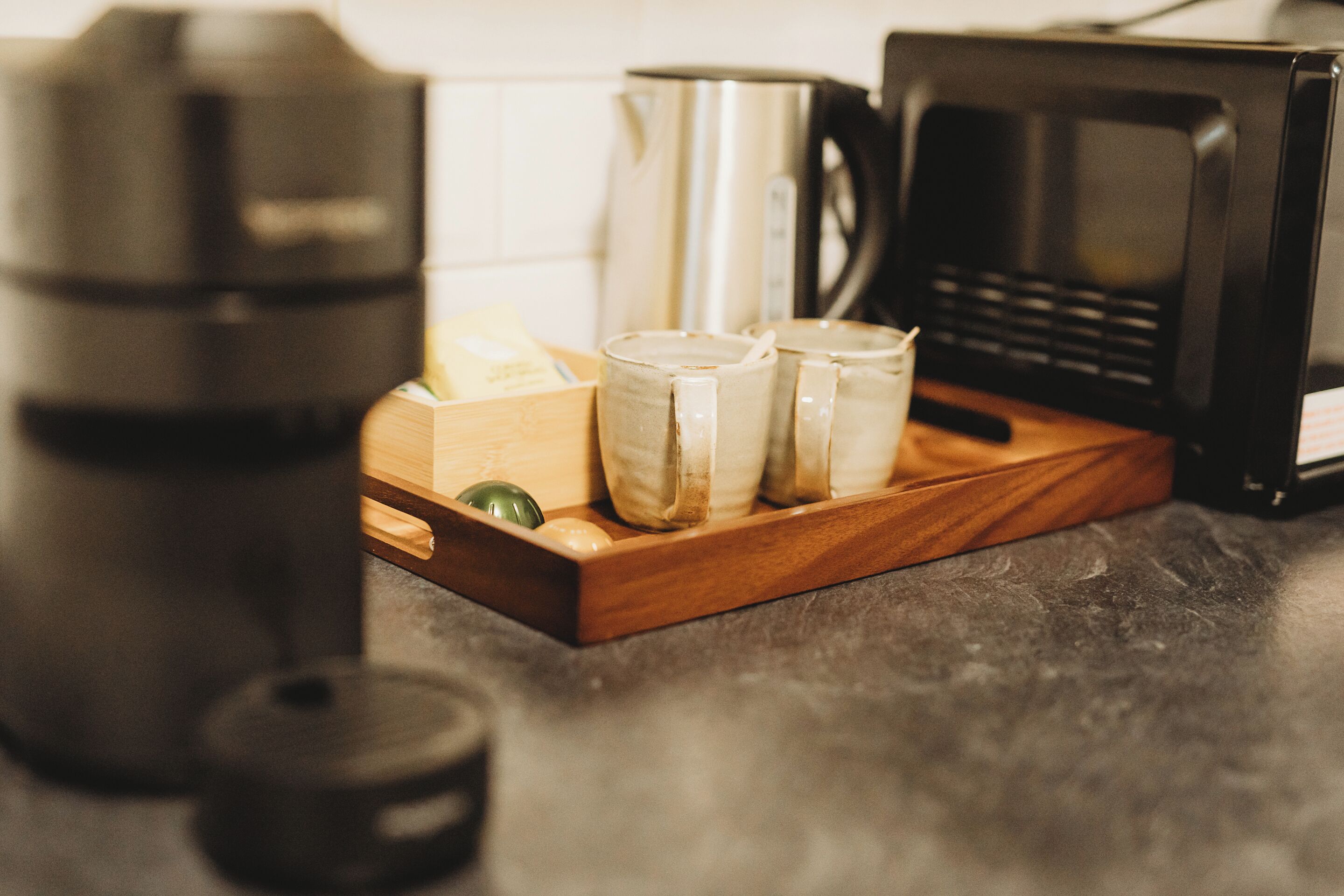 A kitchen counter with a coffee maker, a kettle, and two mugs on a wooden tray.