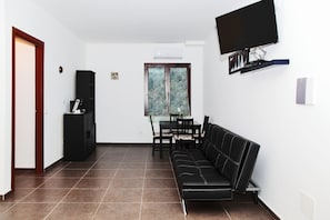 Uma sala de estar com um sofá preto, uma mesa de jantar com cadeiras, um armário preto e uma televisão na parede.
