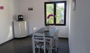 Uma pequena área de refeições com mesa e cadeiras, uma planta em vaso, um refrigerador e uma janela com vista para vegetação.