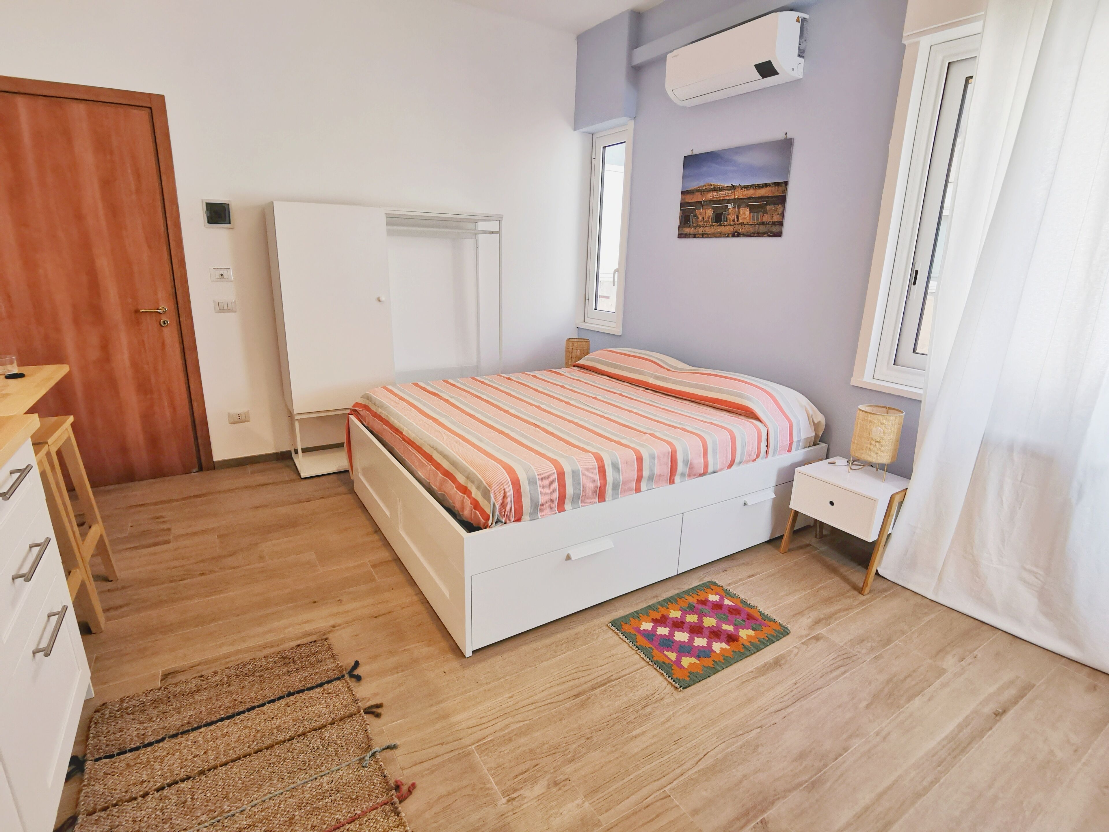 A bedroom with a bed, a bedside table, a lamp, a window with curtains, a wall-mounted air conditioner, and a door.