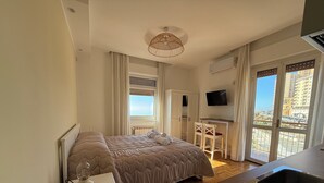 Un dormitorio con cama, escritorio, silla, televisor y balcón con vista a edificios.