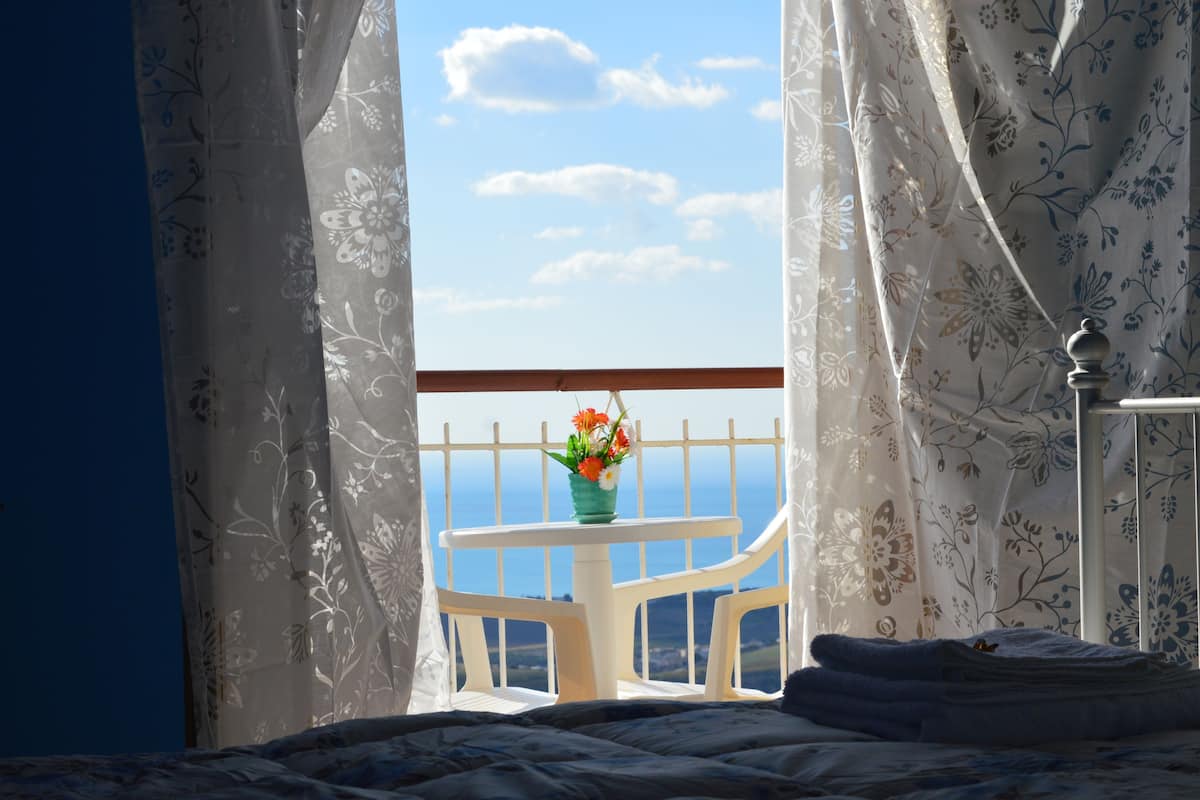 Quarto com uma cama, janela com cortinas, uma mesinha com um vaso de flores e vista para o mar.