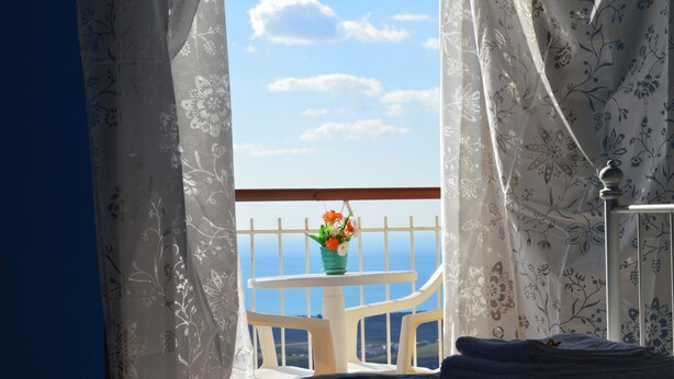 Una habitación con cama, ventana con cortinas, una mesita con un jarrón de flores y vista al mar.