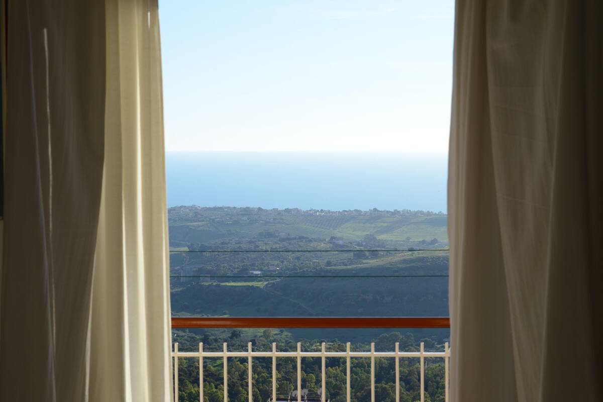 Vista pela janela com cortinas, mostrando uma paisagem com colinas e um horizonte distante.