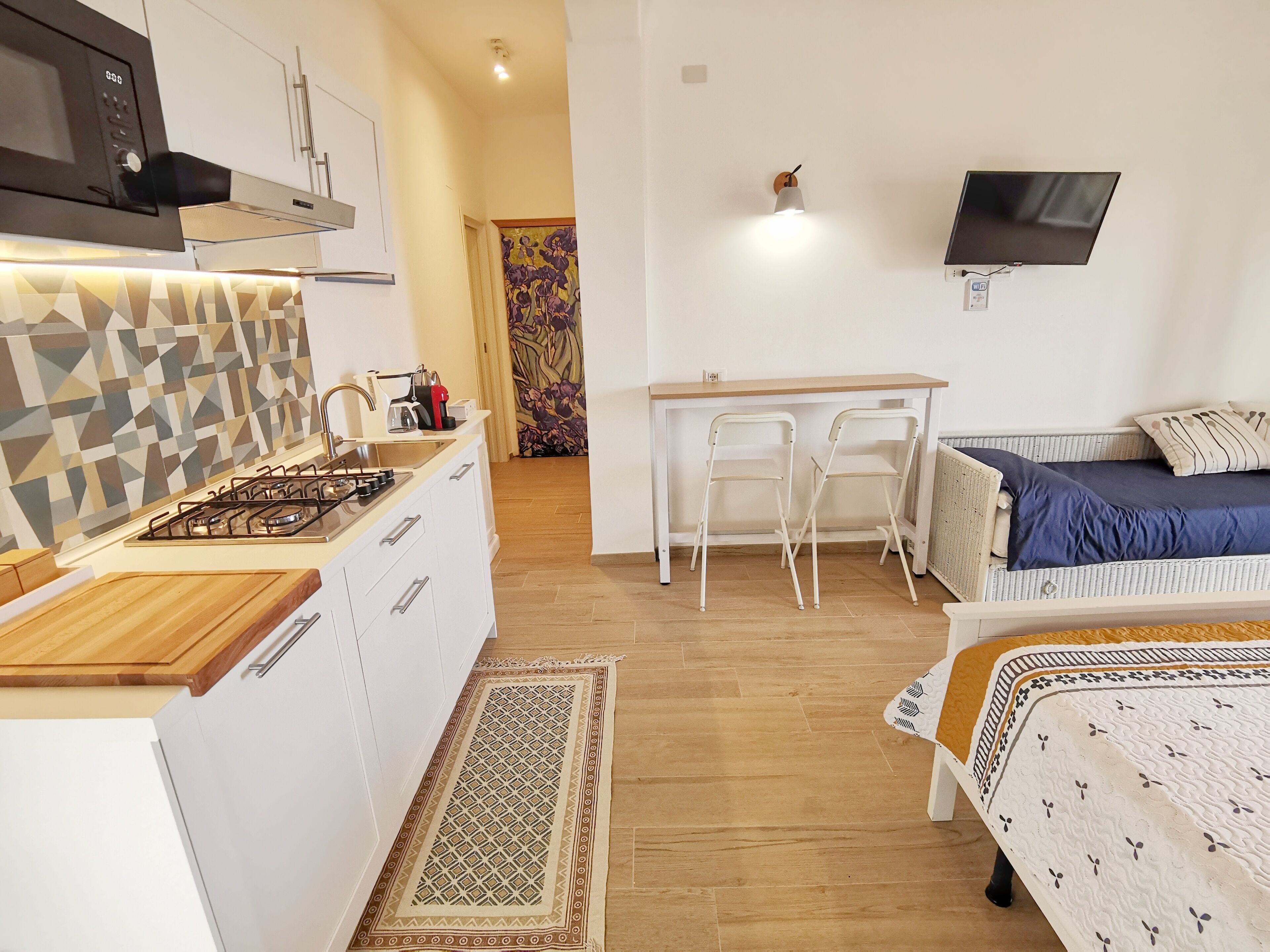 Cocina compacta con estufa a gas, armarios blancos y un salpicadero con diseño. Cuenta con un comedor con mesa y sillas, y una cama con un almohadón azul.