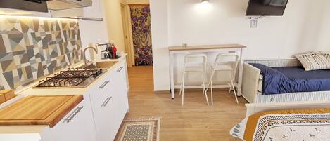 Cocina compacta con estufa a gas, armarios blancos y un salpicadero con diseño. Cuenta con un comedor con mesa y sillas, y una cama con un almohadón azul.