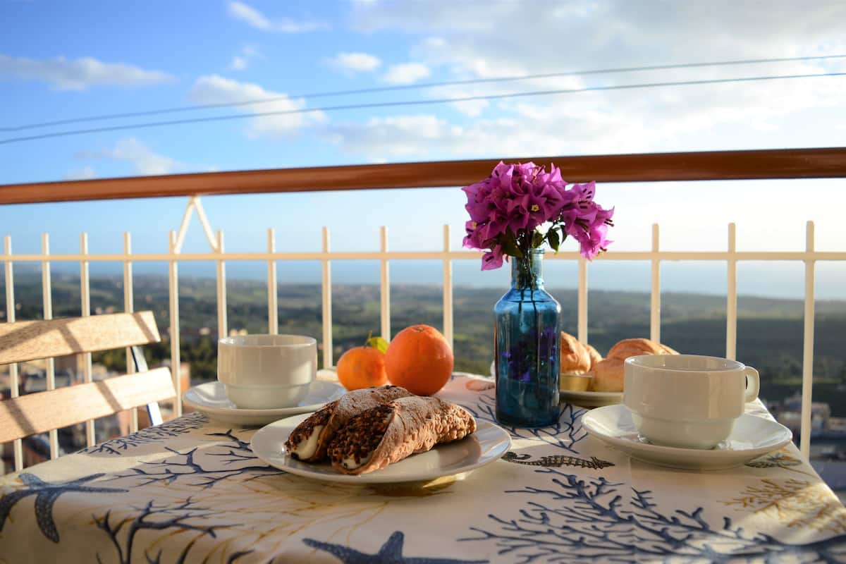 Mesa com doces, um vaso de flores e chávenas numa varanda com vista panorâmica.